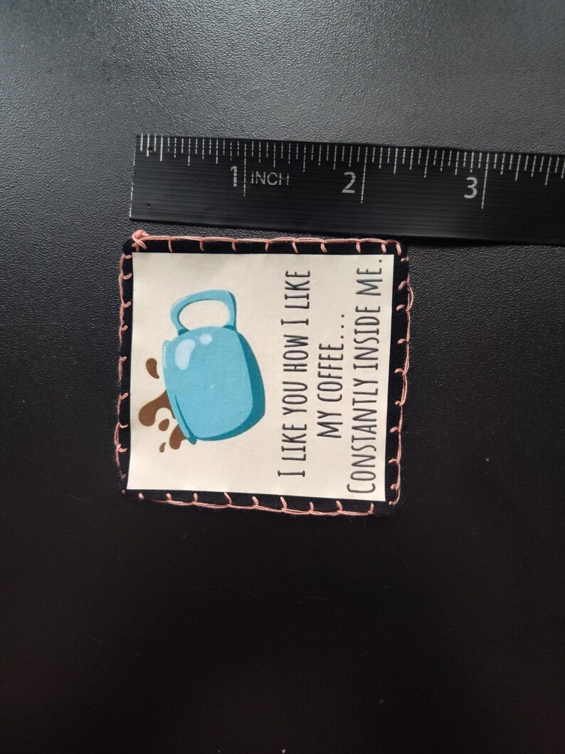 K&ouml;nnte beinhalten: Ein kleines, quadratisches Patch mit schwarzem Rand und rosa N&auml;hten. Das Patch zeigt eine Illustration einer hellblauen Kaffeetasse mit braunen Kaffeespritzern. Der Text lautet: "I LIKE YOU HOW I LIKE MY COFFEE... CONSTANTLY INSIDE ME."