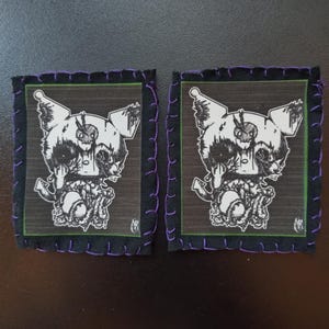 Peut inclure: Deux écussons carrés avec une bordure noire et des coutures violettes. Chaque écusson présente une illustration en noir et blanc d'une créature en forme de crâne avec des tentacules. Le fond est gris foncé avec une bordure verte.