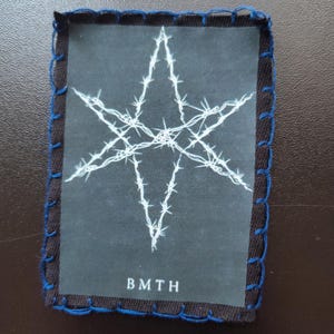 May include: A rectangular patch with a black border and blue stitching. The patch features a white star design made of barbed wire on a dark background. The letters "BMTH" are printed at the bottom of the patch.