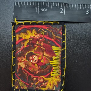 May include: A rectangular patch with a colorful illustration from the movie "ParaNorman." The patch is approximately 2.5 inches long and 2 inches wide, with a black border and yellow stitching. The image features characters in a dynamic, action-oriented scene with the movie title.