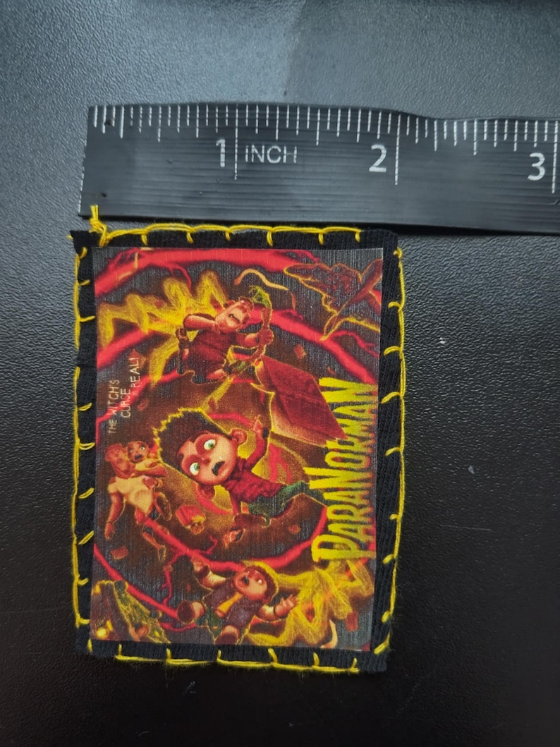 May include: A rectangular patch with a colorful illustration from the movie "ParaNorman." The patch is approximately 2.5 inches long and 2 inches wide, with a black border and yellow stitching. The image features characters in a dynamic, action-oriented scene with the movie title.