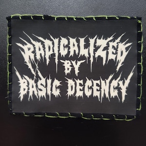 May include: A black rectangular patch featuring the white text "RADICALIZED BY BASIC DECENCY" in a jagged font. The patch is edged with green stitching.