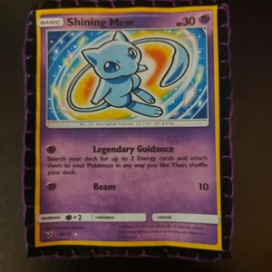 May include: A Shining Mew Pokémon card with a purple border and a light blue Mew character illustration. The card has text including "Legendary Guidance" and "Beam". The card is sewn onto a black fabric with purple stitching.