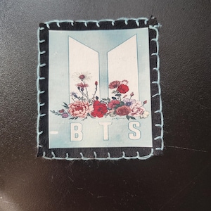 May include: A square patch with a black border and light blue stitching. The patch features the letters "BTS" in white, with a floral arrangement of red, pink, and purple flowers. The background is light blue.