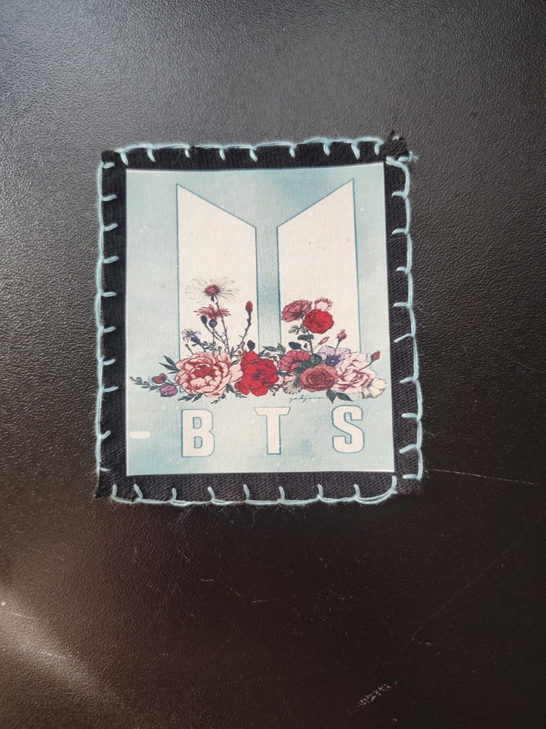 Może przedstawiać: Kwadratowa naszywka z czarną obw&oacute;dką i jasnoniebieskimi przeszyciami. Naszywka zawiera białe litery "BTS" z kwiatowym układem czerwonych, r&oacute;żowych i fioletowych kwiat&oacute;w. Tło jest jasnoniebieskie.