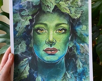 Puede incluir: Una impresión que representa a una mujer con piel verde y ojos amarillos, adornada con una corona de hojas. La obra de arte utiliza tonos de verde y azul, con detalles de hojas y un enfoque en la cara. La impresión es sostenida por una mano.