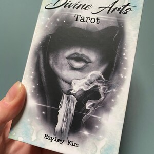 Puede incluir: La caja del mazo de tarot "The Divine Arts Tarot" presenta una ilustraci&oacute;n en blanco y negro de una figura encapuchada con una vela y humo. El texto "The Divine Arts Tarot" y "Hayley Kim" est&aacute; impreso en la caja.
