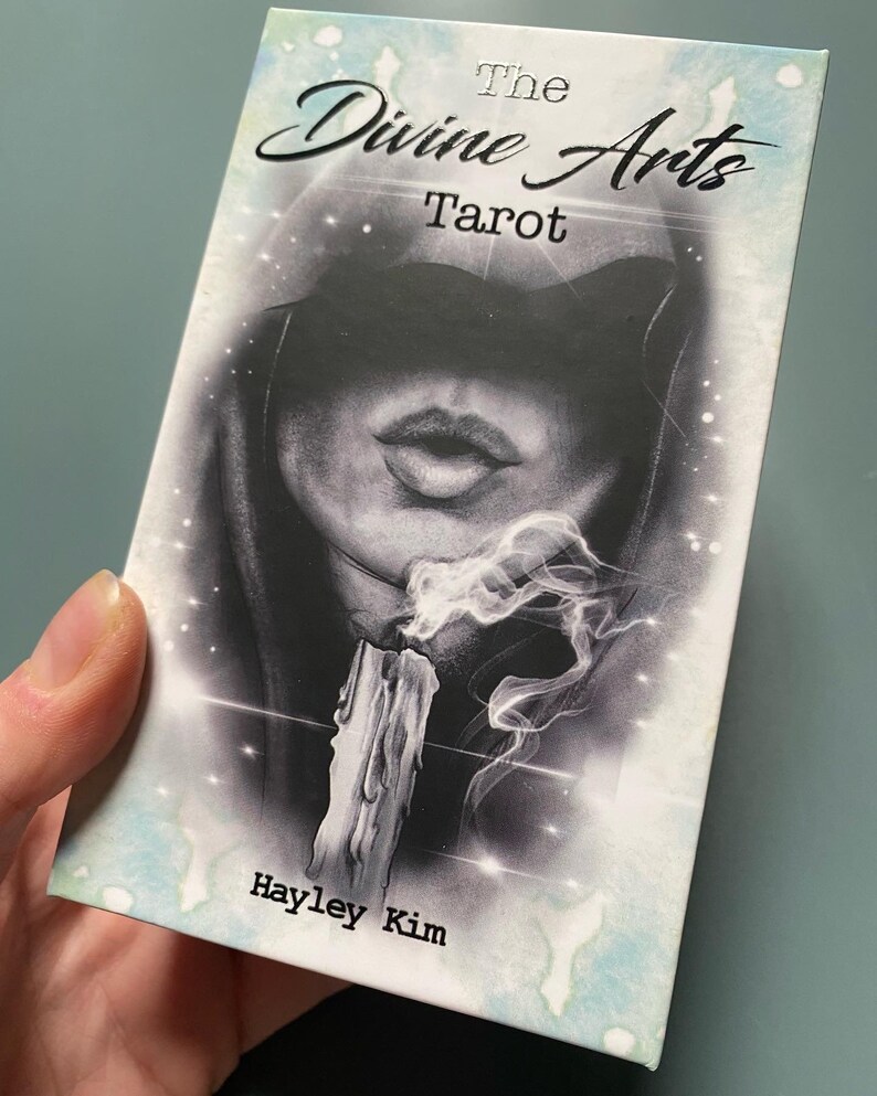 Puede incluir: La caja del mazo de tarot "The Divine Arts Tarot" presenta una ilustraci&oacute;n en blanco y negro de una figura encapuchada con una vela y humo. El texto "The Divine Arts Tarot" y "Hayley Kim" est&aacute; impreso en la caja.