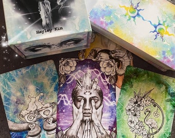 May include: A set of tarot cards with watercolor illustrations. The cards feature a variety of imagery, including a flamingo, a crystal, and a pair of hands. The box for the cards is white with a watercolor design and the text "The Divine Tarot" and "Hayley Kim".