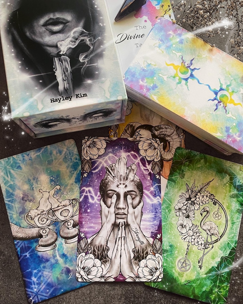 Puede incluir: Un juego de cartas de tarot con ilustraciones de acuarela. Las cartas presentan una variedad de im&aacute;genes, incluyendo un flamenco, un cristal y un par de manos. La caja de las cartas es blanca con un dise&ntilde;o de acuarela y el texto "The Divine Tarot" y "Hayley Kim".