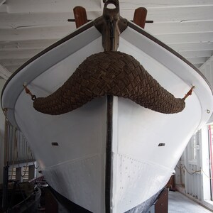 Op de afbeelding: Een witte boot met een zwarte bodem is tentoongesteld. De boot heeft een touw en geweven materiaal dat lijkt op een snor.