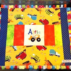 Può includere: Una coperta per bambini personalizzata a tema costruzioni. La coperta ha uno sfondo giallo con vari veicoli da cantiere. Il pannello centrale mostra il nome "Alfie" e un'illustrazione di un trattore. I bordi sono rifiniti con nastri colorati.