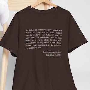 Puede incluir: Una camiseta marrón oscuro con texto blanco. El texto es una cita de la Séptima Enmienda de la Constitución de los Estados Unidos, incluyendo la fecha del 15 de diciembre de 1791. La camiseta se muestra en una percha de madera.