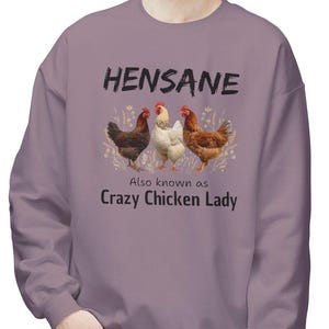 May include: A lilac-colored sweatshirt featuring the text "HENSANE" above an illustration of three chickens. Below the chickens, the text reads "Also known as Crazy Chicken Lady". The chickens are brown, white, and reddish-brown.