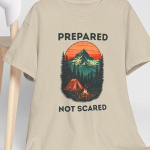 Puede incluir: Camiseta beige con las palabras "PREPARED" y "NOT SCARED" impresas en negro. El gráfico presenta una escena de montaña con una tienda roja, una fogata y árboles, con un atardecer.