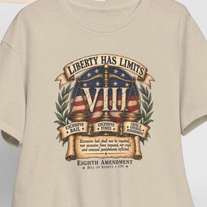May include: A beige t-shirt featuring a graphic of the scales of justice over an American flag. The text reads: LIBERTY HAS LIMITS, VIII, EXCESSIVE BAIL, EXCESSIVE FINES, CRUEL & UNUSUAL PUNISHMENT, EIGHTH AMENDMENT, BILL OF RIGHTS 1791, and legal text.
