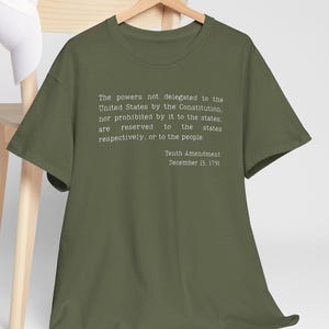 Puede incluir: Camiseta verde oliva con el texto de la Décima Enmienda impreso en blanco. El texto incluye la declaración completa de la enmienda y la fecha del 15 de diciembre de 1791. La camiseta se muestra en una percha de madera.