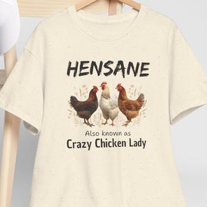 May include: Cream-colored t-shirt with the text "HENSANE" above an illustration of three chickens. Below the chickens, the text reads "Also known as Crazy Chicken Lady".