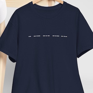 Könnte beinhalten: Marineblaues T-Shirt mit weißen Morsezeichen auf der Brust. Die Morsezeichen ergeben das Wort "LOVE". Das T-Shirt hängt an einem Holzkleiderbügel.