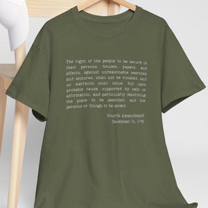Può includere: T-shirt verde oliva con il testo del Quarto Emendamento della Costituzione degli Stati Uniti in bianco. Il testo include la data del 15 dicembre 1791. La maglietta è appesa a una gruccia di legno.