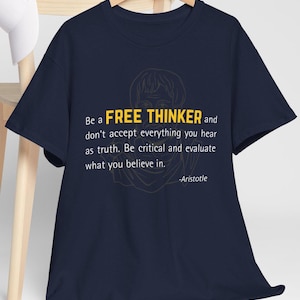 Op de afbeelding: Marineblauw T-shirt met de tekst "Be a FREE THINKER" en een citaat van Aristoteles. Het shirt heeft een gele omtrek van een gezicht. De tekst moedigt kritisch denken en het evalueren van overtuigingen aan.