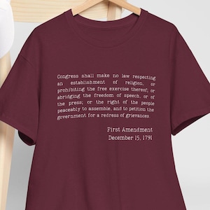 Puede incluir: Una camiseta granate con el texto de la Primera Enmienda impreso en blanco. El texto incluye el derecho a la libertad de expresión, religión y prensa. La fecha del 15 de diciembre de 1791 también está impresa en la camiseta.