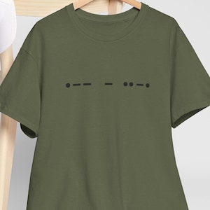 May include: An olive green t-shirt featuring a black Morse code design. The design is made up of dots and dashes, forming a minimalist pattern. The t-shirt is displayed on a wooden hanger.
