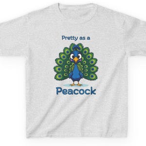 Op de afbeelding: Lichtgrijs t-shirt met een cartoon pauw ontwerp. De pauw is blauw met groene en gele staartveren. De tekst "Pretty as a Peacock" en "Peacock" zijn blauw.