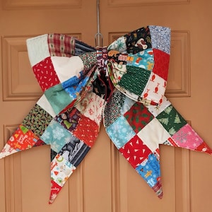 May include: A large, handmade quilted bow in a patchwork design, perfect for holiday decor. The bow features a variety of fabrics in red, green, white, and blue, with festive patterns like snowflakes, reindeer, and Christmas trees. It is hanging on a door.
