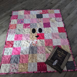 May include: A handmade patchwork quilt in shades of pink, white, and gray, featuring a whimsical ghost face design. The quilt is rectangular with a pink border and a book titled "The Little Ghost Who Was a Quilt" is placed on top.