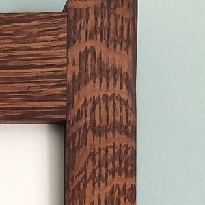 May include: Close-up of a wooden picture frame corner. The frame is made of dark brown wood with a prominent grain pattern. The frame surrounds a white surface, likely a canvas or paper, against a light blue wall.