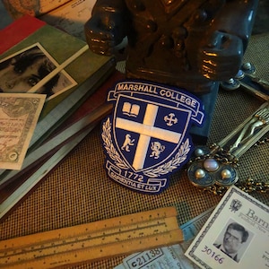 Puede incluir: Un parche azul y blanco de Marshall College con el texto "Marshall College" y "Sapientia Et Lux". El parche está rodeado de libros, una regla y otros objetos antiguos.