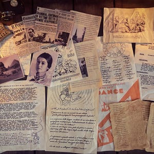 May include: A collection of vintage ephemera, including newspapers, photographs, and handwritten documents, arranged on a wooden surface. The items have a sepia tone, suggesting age, and include text in multiple languages. A vintage camera and dominoes are also present.