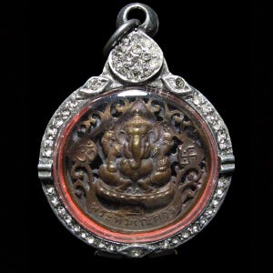 Puede incluir: Un colgante detallado que presenta una representación en color bronce de la deidad hindú Ganesha, sentado sobre un loto. El colgante está encerrado en un marco plateado con un centro transparente y un borde rojo. El marco está adornado con pequeños acentos brillantes.