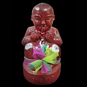 Op de afbeelding: Rood beeldje van een zittende baby met handen bij de mond. Het beeldje heeft witte accenten op de knieën en is versierd met een kleurrijke strik. De basis van het beeldje heeft tekst in een vreemde taal.