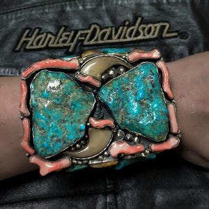 May include: A rectangular belt buckle featuring two large, irregular turquoise stones, two curved animal claws, and multiple pieces of smooth, light pink coral set in a silver-toned metal frame with small decorative silver beads and star accents.