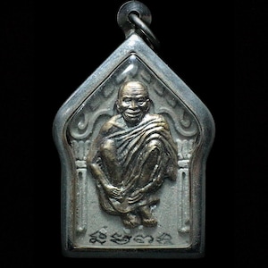 May include: A metallic pendant featuring a bronze-toned relief of a seated figure with a shaved head, draped in robes. The figure is encased in a silver-colored, pointed-arch frame with a top loop for hanging. The base displays embossed script characters.