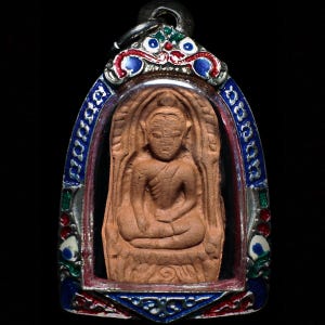 Può includere: Un ciondolo con una figura di Buddha seduto in una cornice protettiva. Il Buddha è di colore marrone chiaro e la cornice è decorata con smalto blu, rosso e verde. Il ciondolo ha un'ansa color argento per appenderlo.