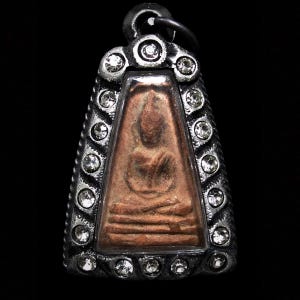 May include: A silver-toned pendant with a detailed, seated Buddha figure in a rectangular frame. The frame is embellished with clear, round crystals. The pendant has a loop at the top for a chain and is set against a black background.