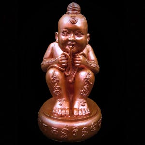 Op de afbeelding: Een bronzen beeld van een zittende, kinderlijke figuur met een knot en gezichtsmarkeringen. De figuur houdt een lang, slangachtig object in zijn mond en heeft inscripties op zijn benen en basis. Het beeld staat op een ronde basis tegen een zwarte achtergrond.
