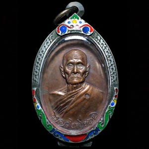 May include: A bronze-colored pendant with an oval shape, featuring a detailed relief of a man's face. The pendant has a decorative border with colorful enamel accents in red, blue, and green. A small metal loop is at the top for hanging.