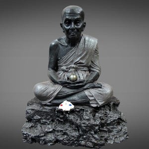 Könnte beinhalten: Eine sitzende Statue eines Mönchs in meditativer Haltung, der eine Kugel hält. Die Statue ist dunkelgrau mit silbernen Akzenten und steht auf einem strukturierten schwarzen Sockel. Ein kleines, weißes Objekt mit roten und blauen Details liegt auf dem Sockel.