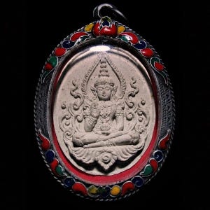 Könnte beinhalten: Ein ovaler Anhänger mit einer sitzenden Buddha-Figur in einem detaillierten, hellfarbigen Relief. Der Anhänger ist in einen silbernen Rahmen mit einem farbenfrohen Emaille-Rand mit roten, blauen, grünen und gelben Akzenten gefasst. Der Hintergrund ist schwarz.