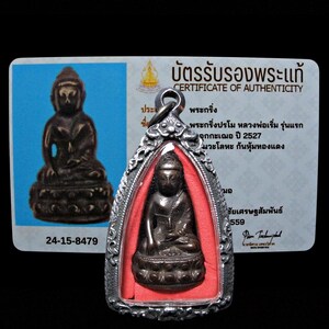 May include: A bronze Buddha amulet pendant with a detailed design, encased in a silver frame. The pendant is displayed with a certificate of authenticity, featuring text in Thai and English. The background is black.