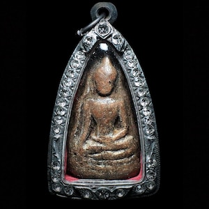 May include: A brown, textured, seated Buddha figure encased in a pointed, silver-toned metal pendant frame. The frame is decorated with numerous small, clear, sparkling stones along its entire perimeter and features a circular bail at the top.