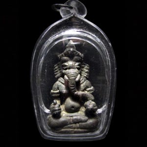May include: A small, dark-colored statue of the Hindu deity Ganesha, encased in a clear, protective plastic pendant. The statue features intricate details, including an elephant head and multiple arms. The pendant is suspended from a small loop.
