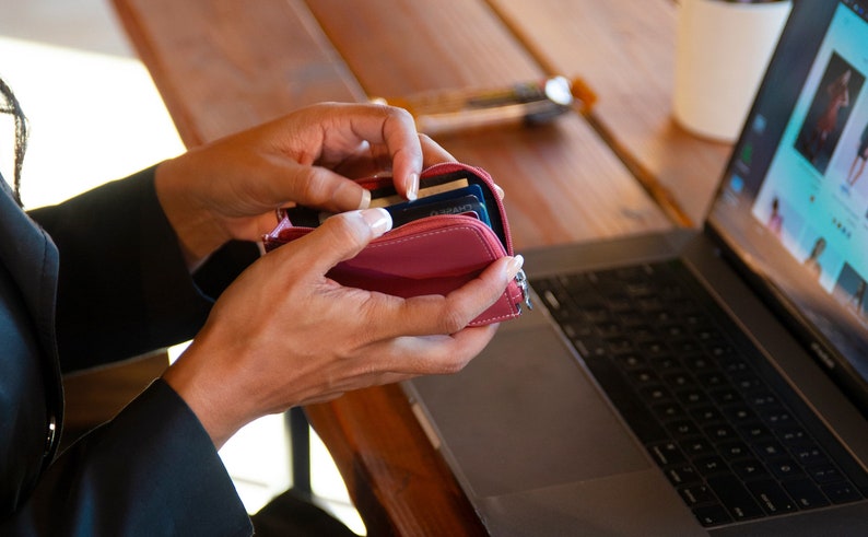May include: A person's hands holding a pink leather wallet open, revealing multiple credit cards inside. The wallet is on a wooden table next to a laptop computer.