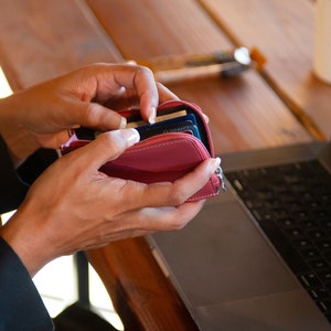May include: A small, red leather wallet with a zipper closure is being opened to reveal credit cards. The wallet is held in front of a laptop on a wooden table. The laptop screen displays a website.