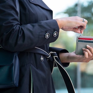 May include: A gray leather wallet with a zipper closure, held open to reveal credit cards. The wallet is being held by a person wearing a black coat and carrying a dark blue purse. The purse has a silver clasp.