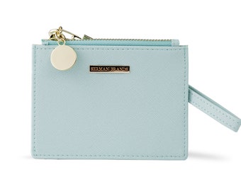 May include: A pale blue wallet with a gold zip and a matching wrist strap. The wallet features a gold circular charm and a gold rectangular label reading "SERMAN BRANDS".